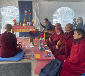 We see teachers dressed in monastic robes performing a ground blessing ceremony with Dzogchen Ponlop Rinpoche in suit at the head throwing flowers and the Gandhara Buddha image on the background.