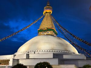 The white big stupa in Boudha, Nepal, with a dark sky but light shining on the stupa and eyes of the Buddha clearly visible.